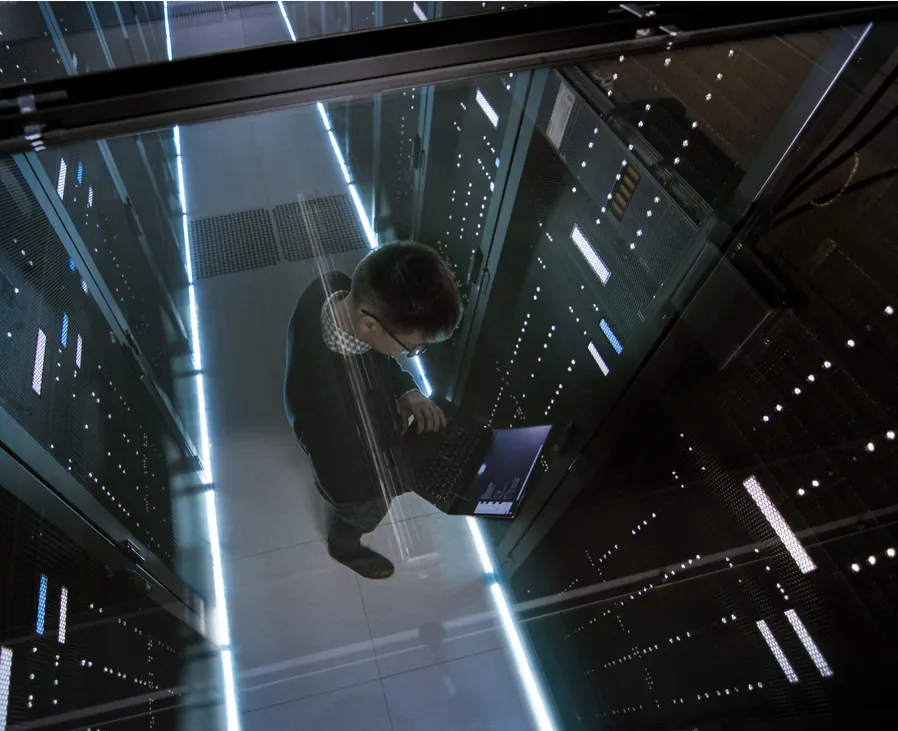 Aerial view through a glass window of an IT engineer working on a laptop within a data center full of active servers.